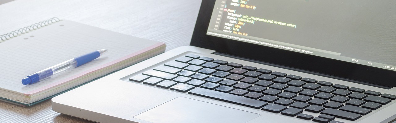 Photo of an open Macbook laptop with code on the screen and an open notepad and pen next to it.
