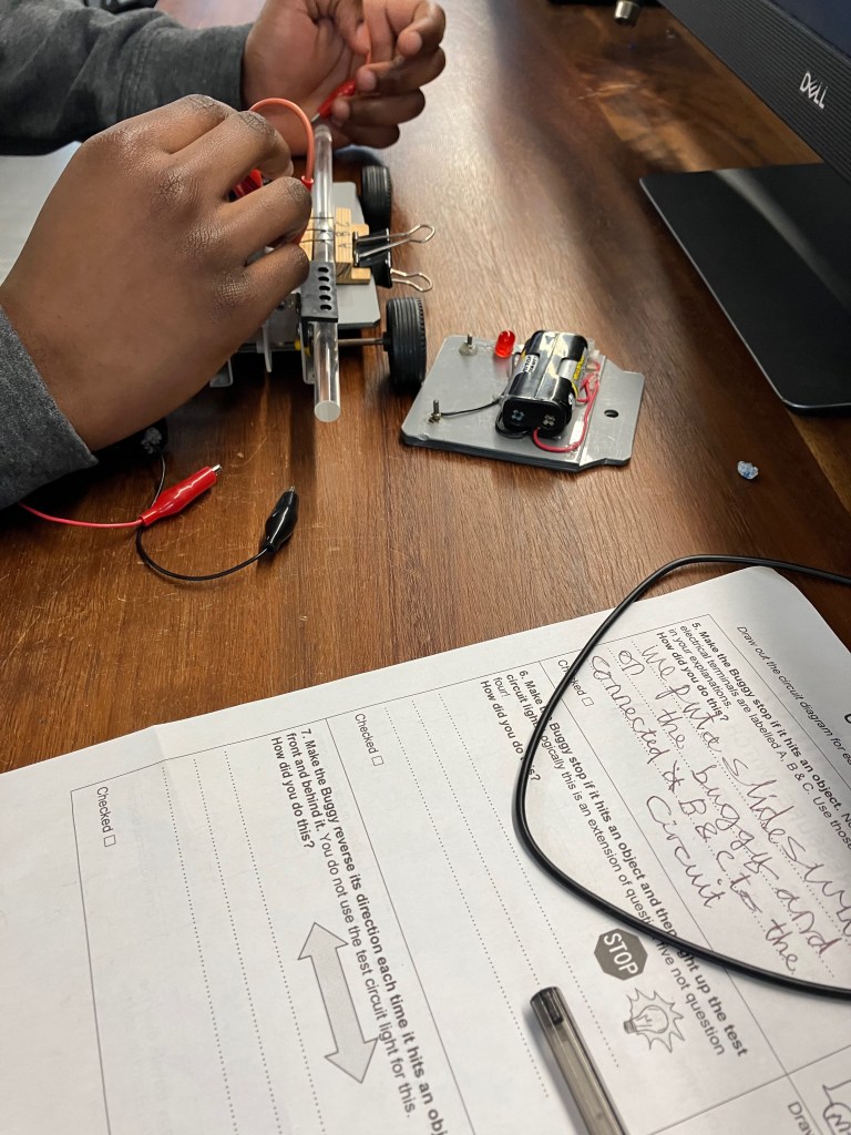A young person's hands holding some wires and connecting them in the correct place on their buggy. On the right, the ever present and very important documentation - the students fill in what they've done. An important skill in engineering or computer science.