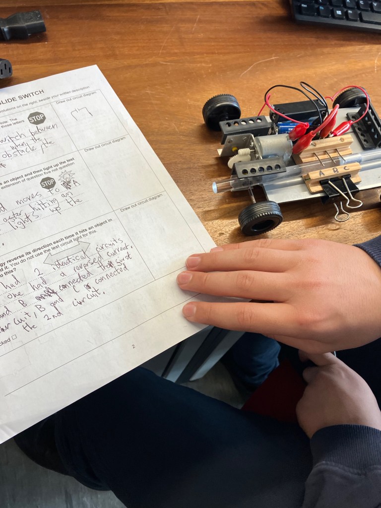 A young person's hand holding a bit of paper with some writing on it, next to a small wheeled buggy with a battery and wired connectors. The person is checking that they've wired it correctly and making notes.