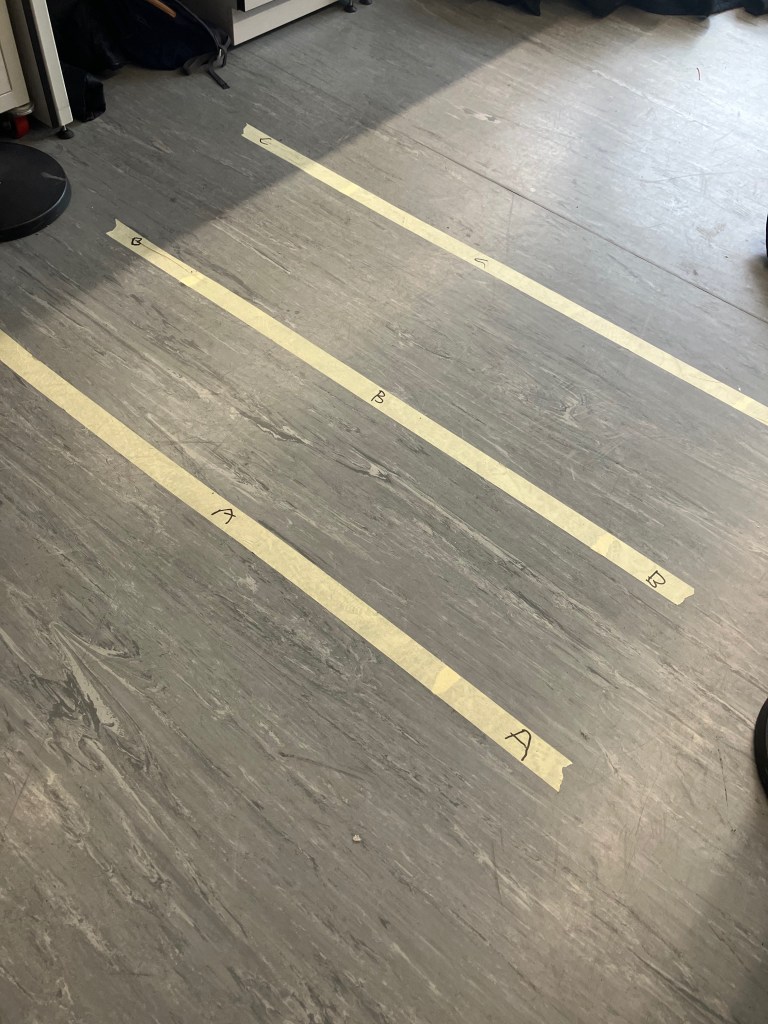 An ordinary grey linoleum lab floor but with three strips of masking tape laid out and labelled A, B and C. A and B are 40 centimetres apart, B and C are 50 centimetres apart. The class are programming their wheeled buggies to move automatically from A to B, then pause, then from B to C and pause, then reverse the process.
